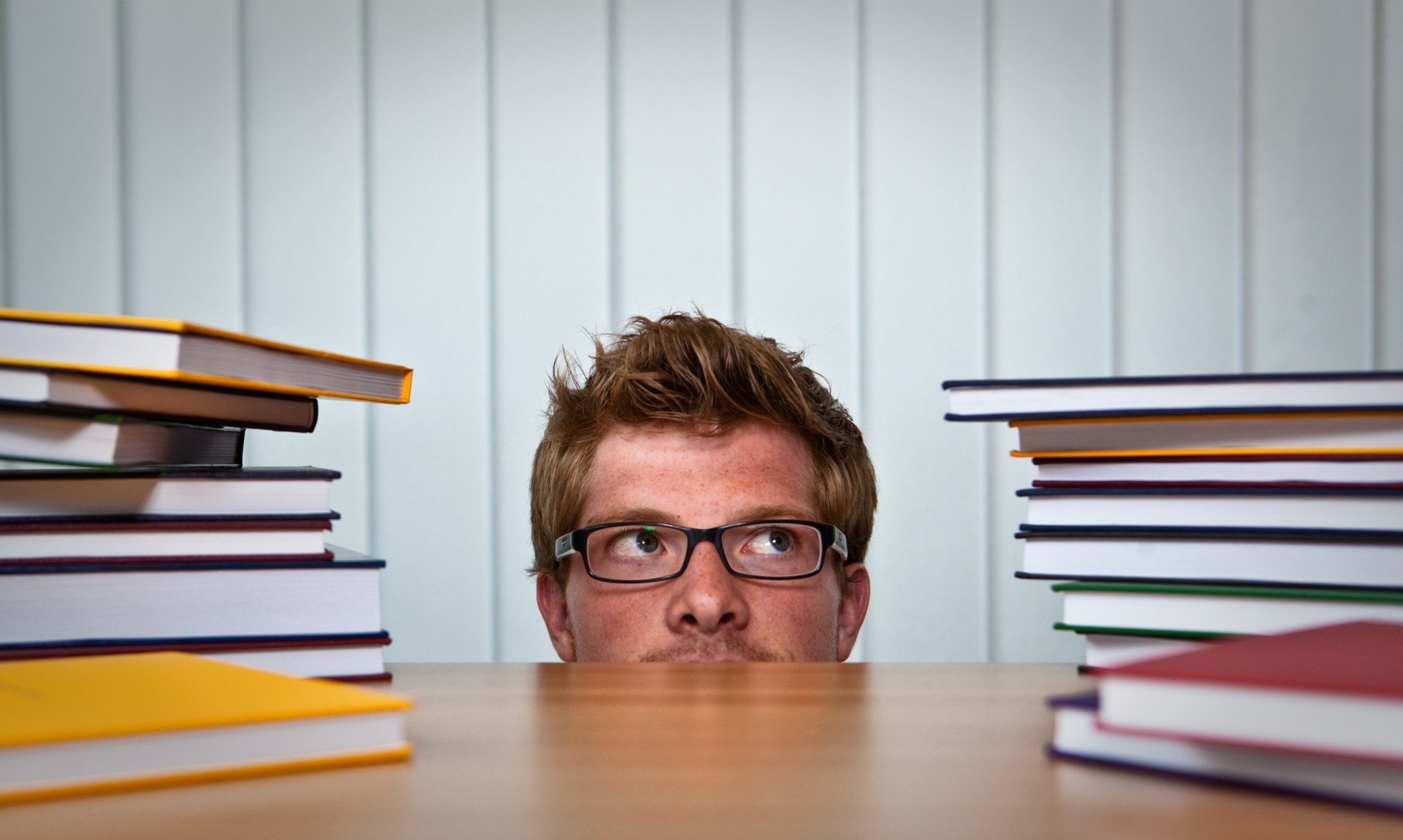 Im Vordergrund ist ein Tisch auf dem links und rechts jeweils hohe Bücherstapel sind. Hinter dem Tisch und zwischen den Büchern ragt der großteil eines Kopfes hervor.